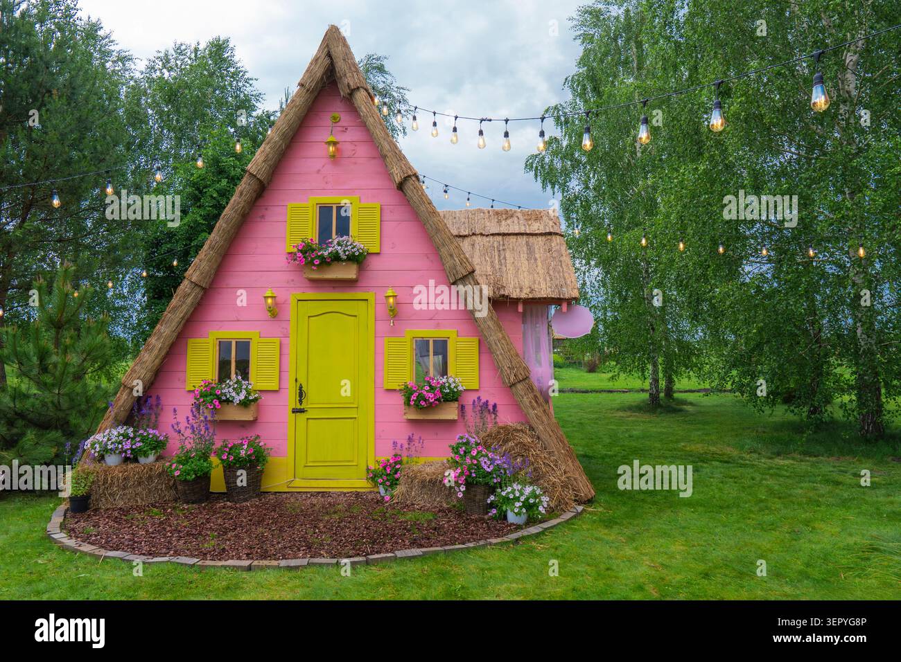 Incantevole decorazione di cottage rosa in un parco pubblico. Una stravagante zona fotografica con tetto in paglia, fiori e luci a corda su un prato sotto il cielo nuvoloso Foto Stock