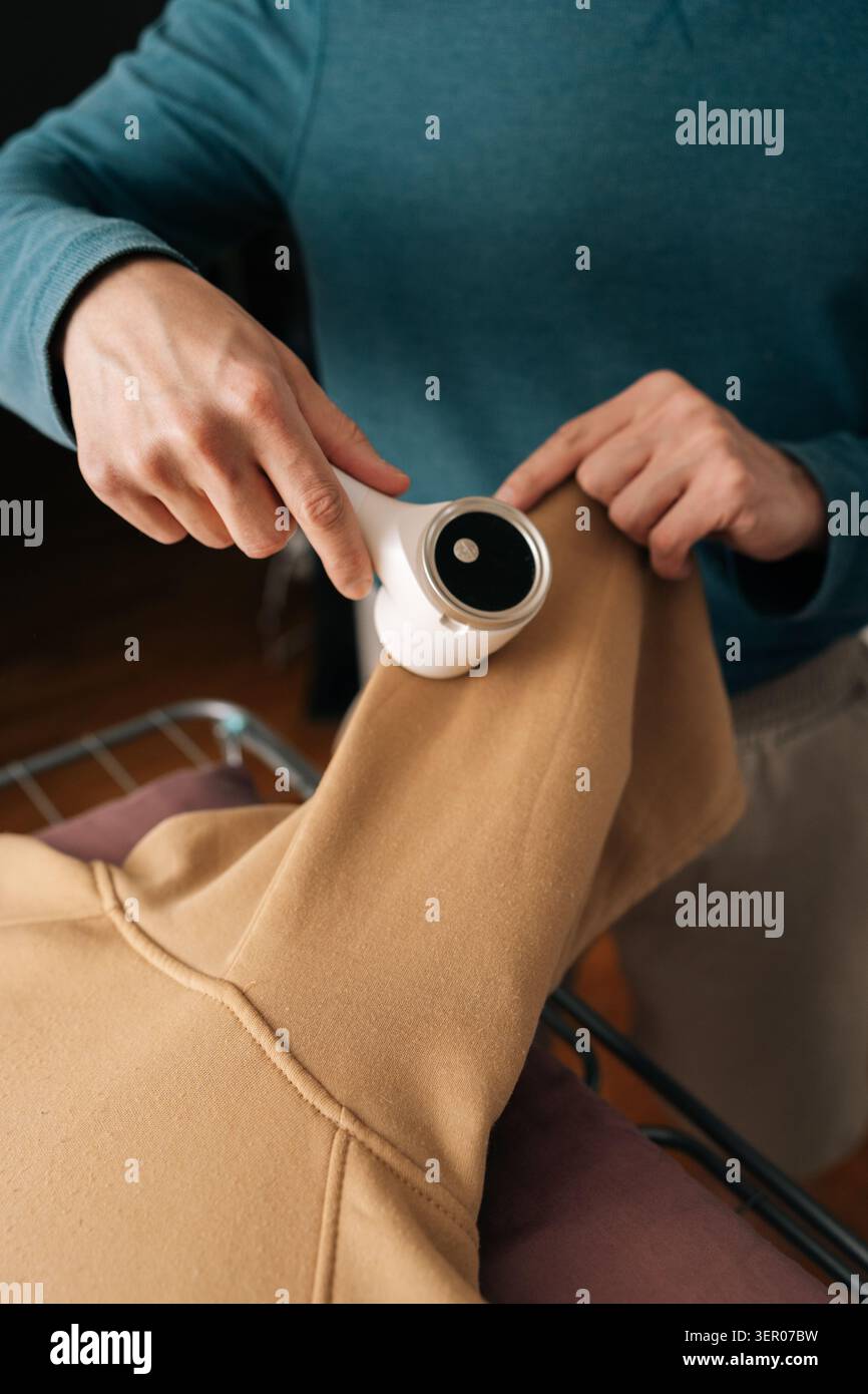 Primo piano verticale di una persona che utilizza un rasoio elettrico per tessuti, rimuovendo la lanugine dalla manica beige della felpa per la cura degli indumenti e un abbigliamento rinnovato Foto Stock