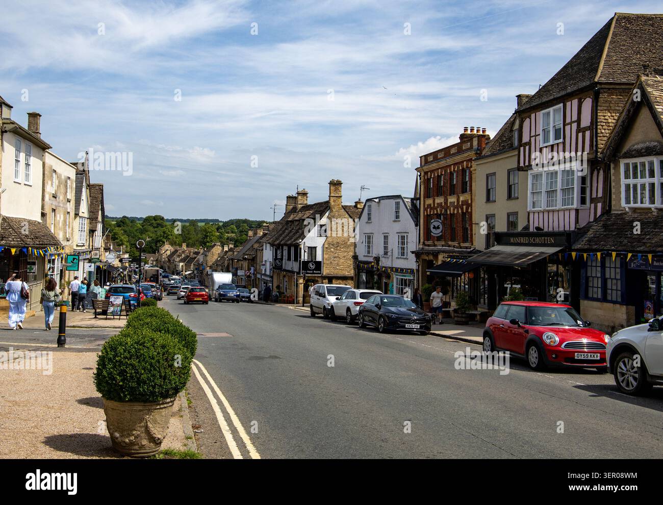 Un'affascinante scena stradale nella storica città inglese di Burford caratterizzata da una tradizionale architettura in pietra, auto che guidano su strada e pedoni Foto Stock