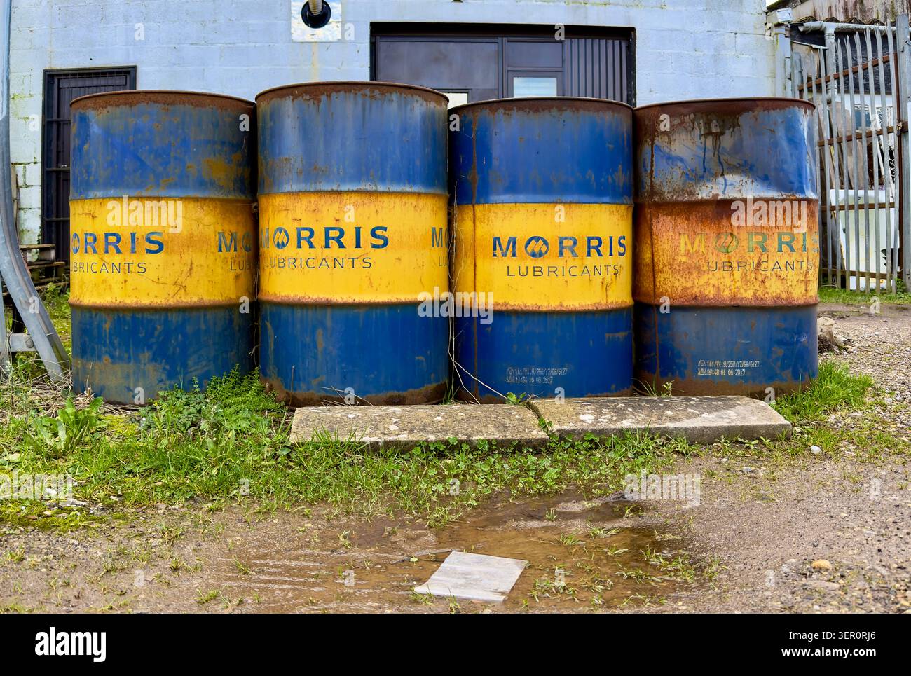Quattro fusti di olio blu e giallo Rusty Morris Lubricants, situati all'esterno di un edificio industriale Foto Stock