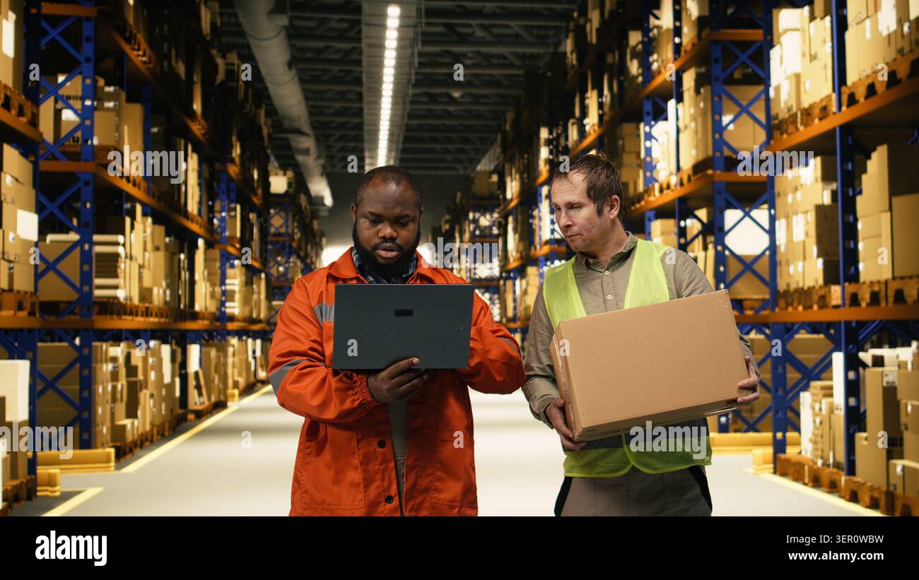 Due colleghi del magazzino organizzano pallet e pacchi con un computer portatile, controllando le informazioni di controllo dell'inventario in un deposito industriale. Il lavoro di squadra migliora il flusso di lavoro operativo e i processi di e-commerce. Foto Stock