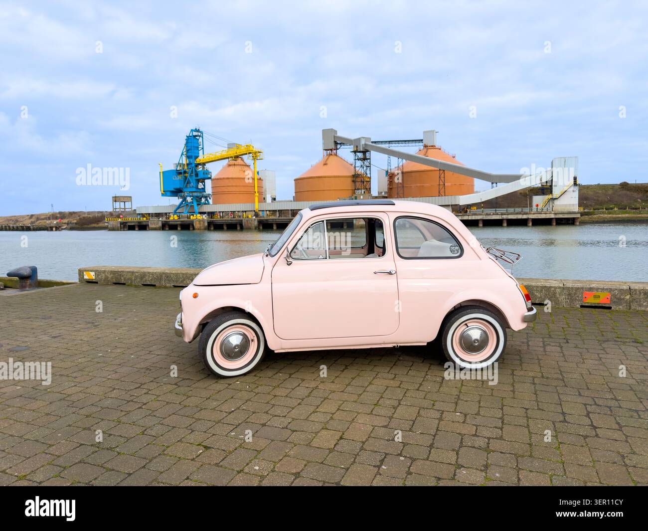 Una classica fiat 500 rosa parcheggiata in un porto industriale con gru e silos sullo sfondo - Immagine stock catturata con smartphone