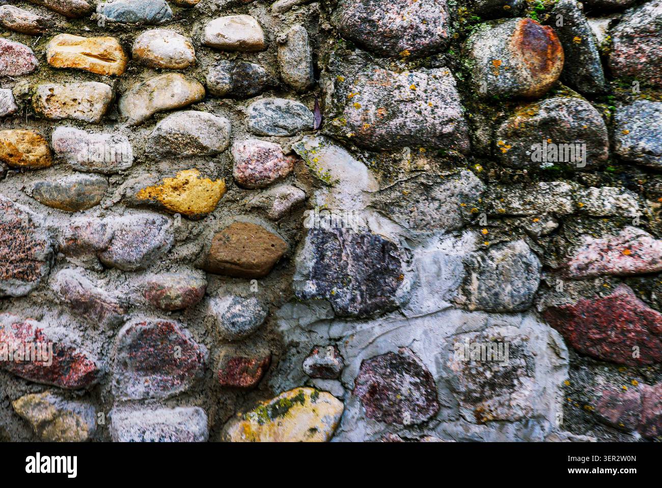 Muschio e lichene si aggrappano a superfici irregolari. Le pietre grigie, marroni e ocra si fondono con la Malta usurata. Il muro suggerisce età, resilienza e resistenza silenziosa Foto Stock