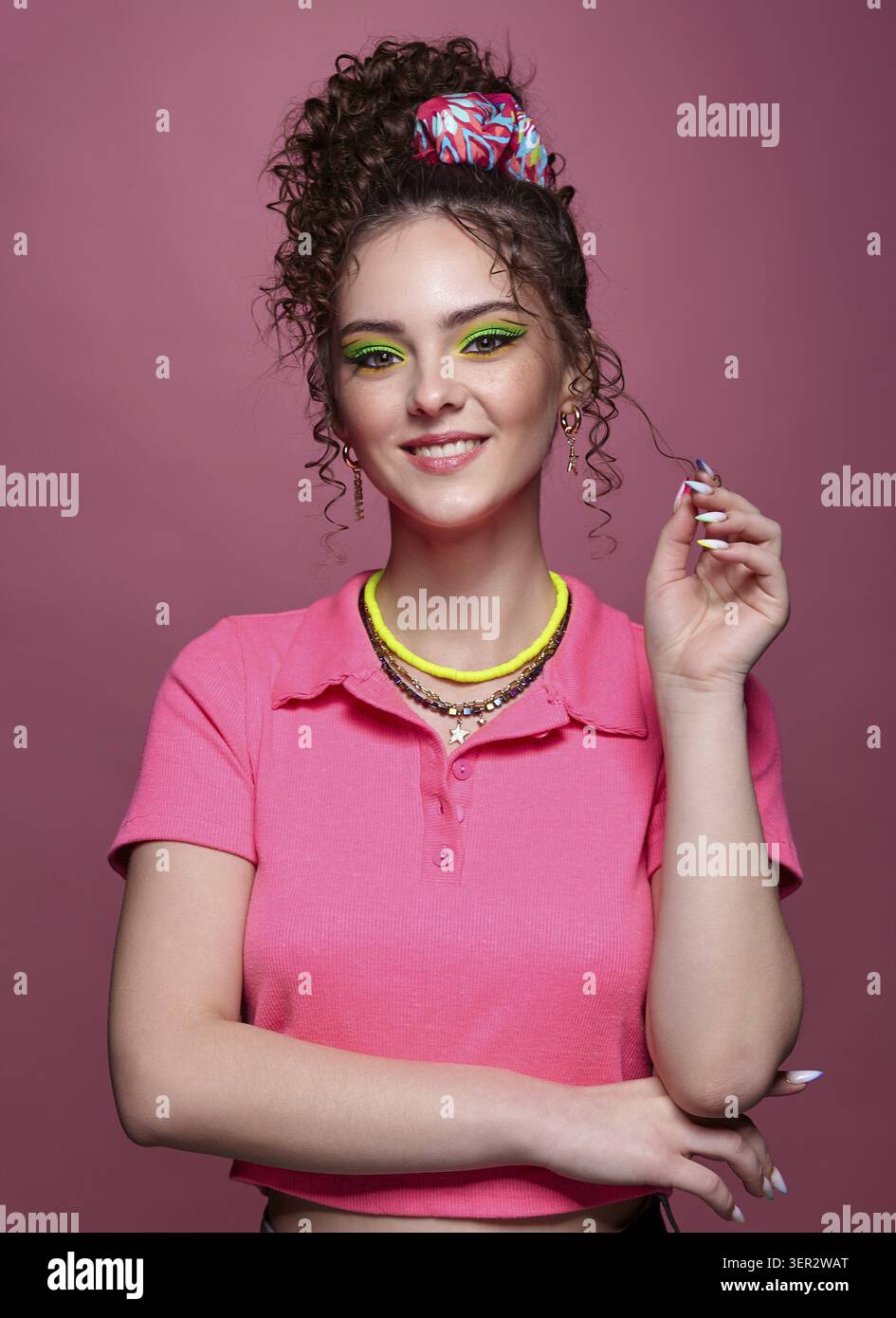 Ritratto di una giovane donna sorridente su sfondo rosa. Donna con insoliti occhi verdi ombre trucco, capelli ricci e orecchini Foto Stock