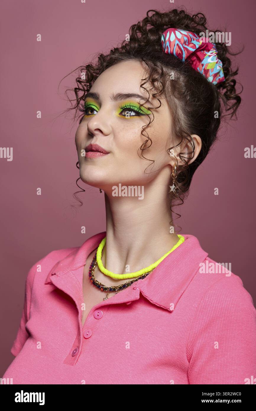 Ritratto di una giovane donna sorridente vestita con un abito rosa su sfondo rosa. Donna con insoliti occhi verdi ombre trucco, capelli ricci e orecchini Foto Stock