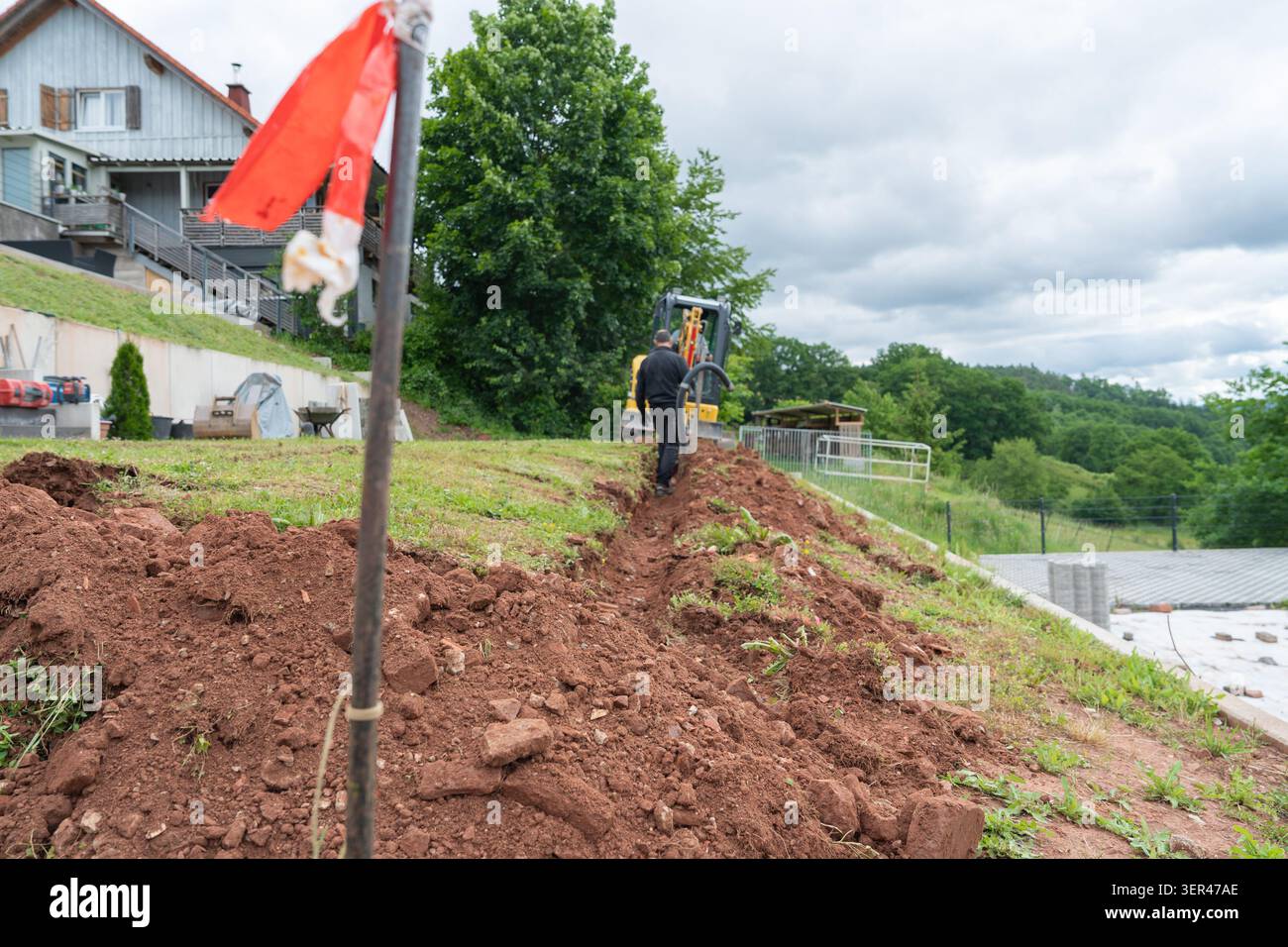 Una lunga trincea aperta si estende su un profilo di collina erboso con una bandiera rossa di sicurezza che segna la zona di scavo. Giardino residenziale Foto Stock