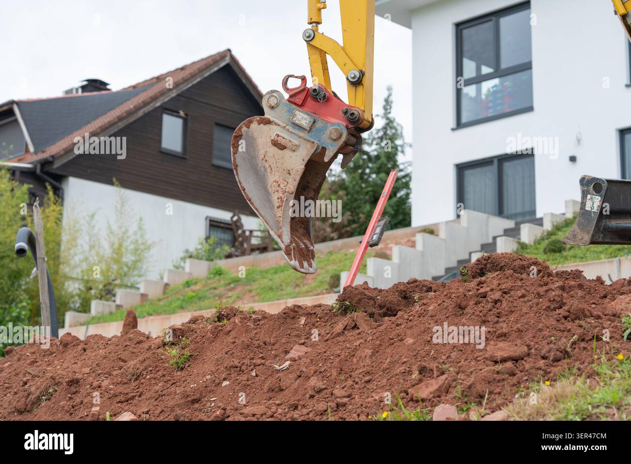 Un mini escavatore giallo esegue lavori in terra e il livellamento del terreno su un ripido pendio sul retro di fronte a una moderna casa residenziale. Architettura paesaggistica Foto Stock