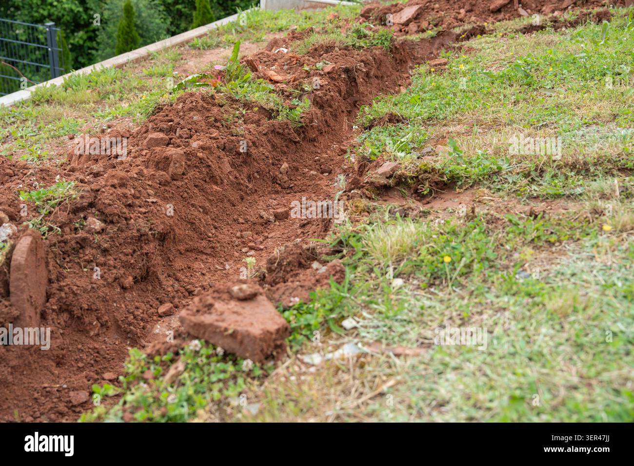 Una trincea lunga e stretta scavata in terreno marrone scuro su un pendio verde erboso per l'installazione di tubi per servizi pubblici residenziali. Aperto Foto Stock