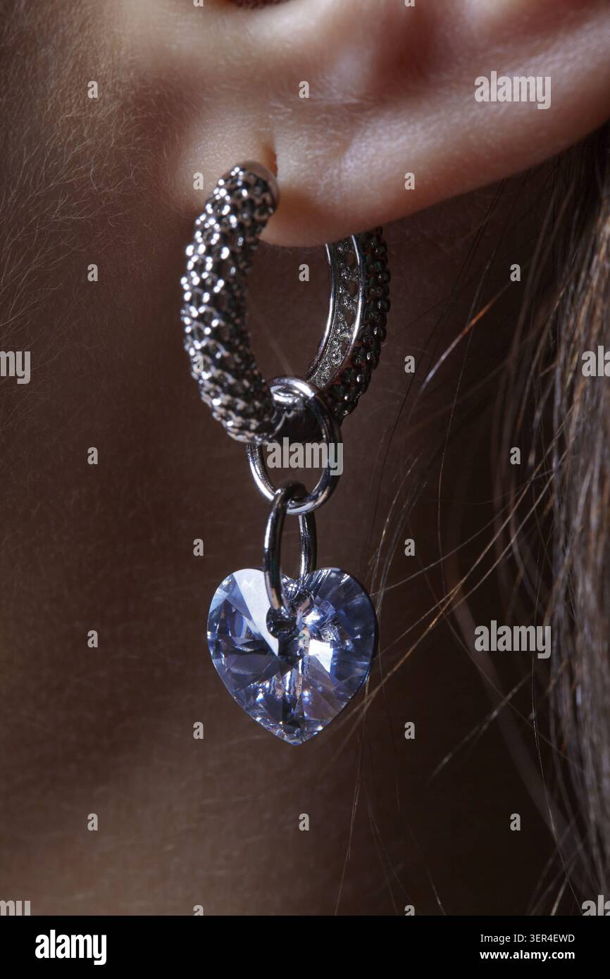 Orecchio umano femminile con orecchino. L'auricolare è composto da un anello in metallo modellato e da un strass blu a forma di cuore Foto Stock