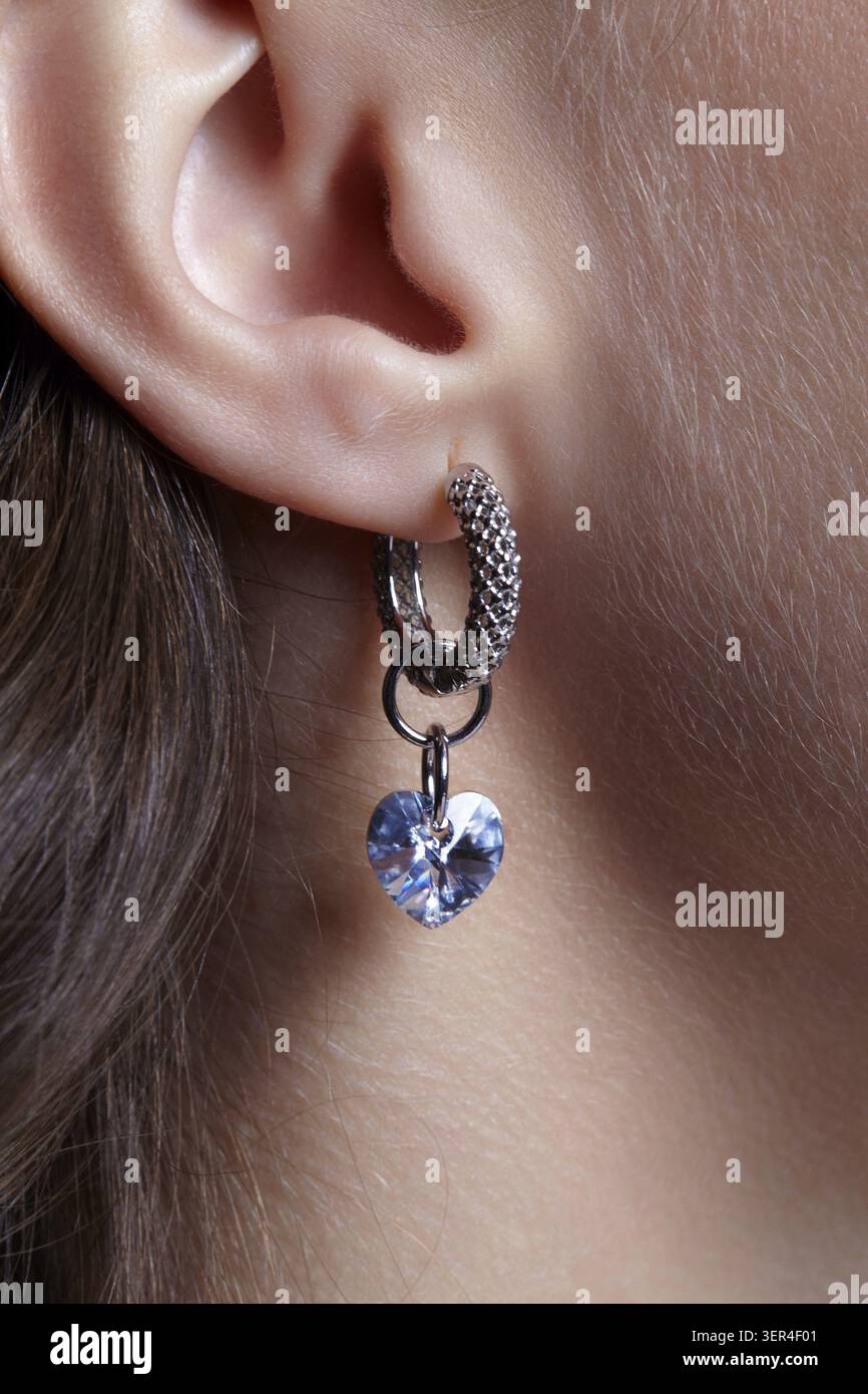 Orecchio umano femminile con orecchino. L'auricolare è composto da un anello in metallo modellato e da un strass blu a forma di cuore Foto Stock