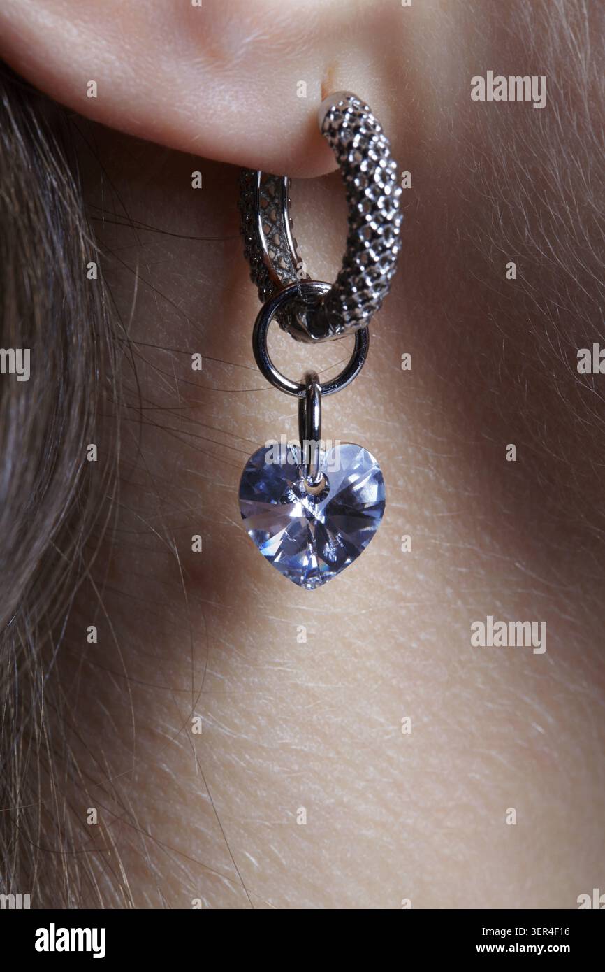 Orecchio umano femminile con orecchino. L'auricolare è composto da un anello in metallo modellato e da un strass blu a forma di cuore Foto Stock