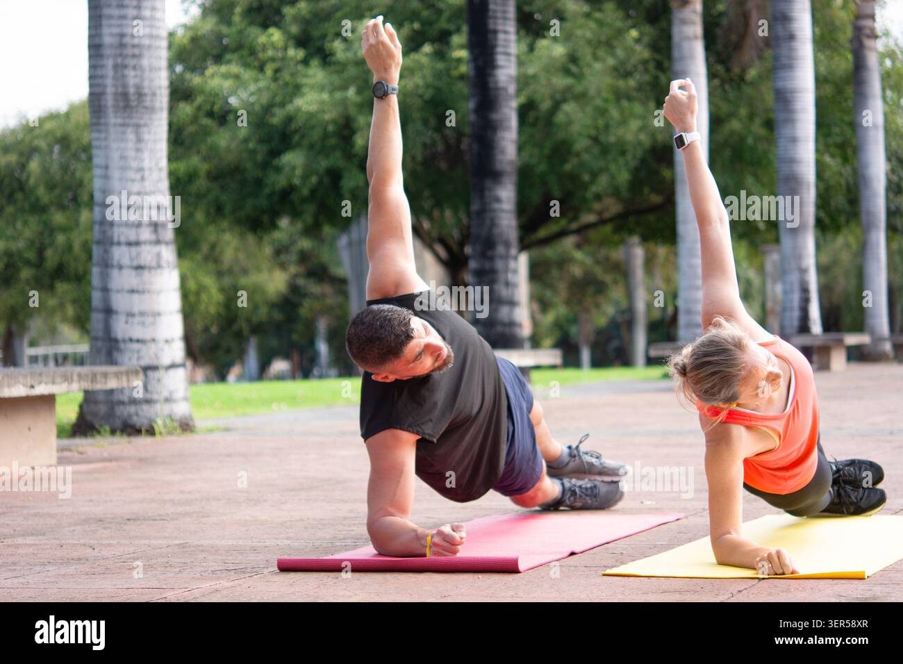 Uomo e donna maturi che praticano esercizi con tavole laterali su tappetini da yoga in un parco soleggiato. Foto Stock