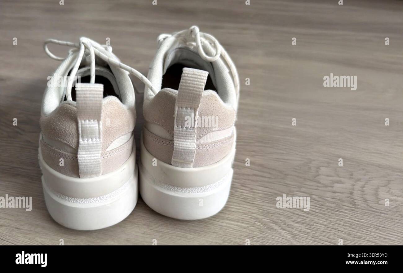 Sneaker atletiche bianche e beige con lacci e linguette sul tallone posizionate su un pavimento in legno chiaro, caratterizzate da un design moderno adatto per l'abbigliamento casual Foto Stock