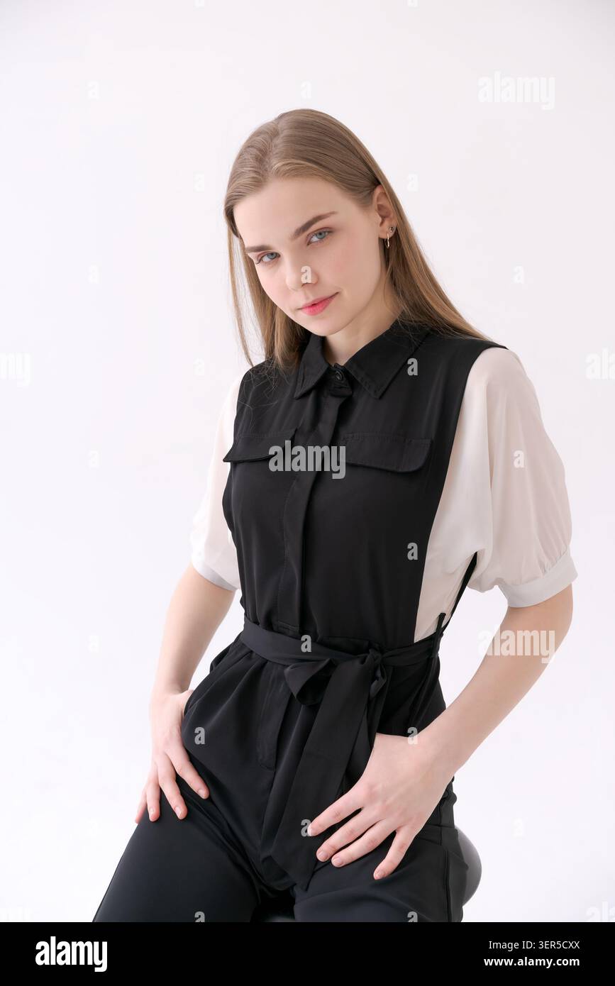 Una giovane donna bionda con le mani sui fianchi in una tuta senza maniche nera sopra una camicetta bianca a maniche di maniche in uno studio Foto Stock