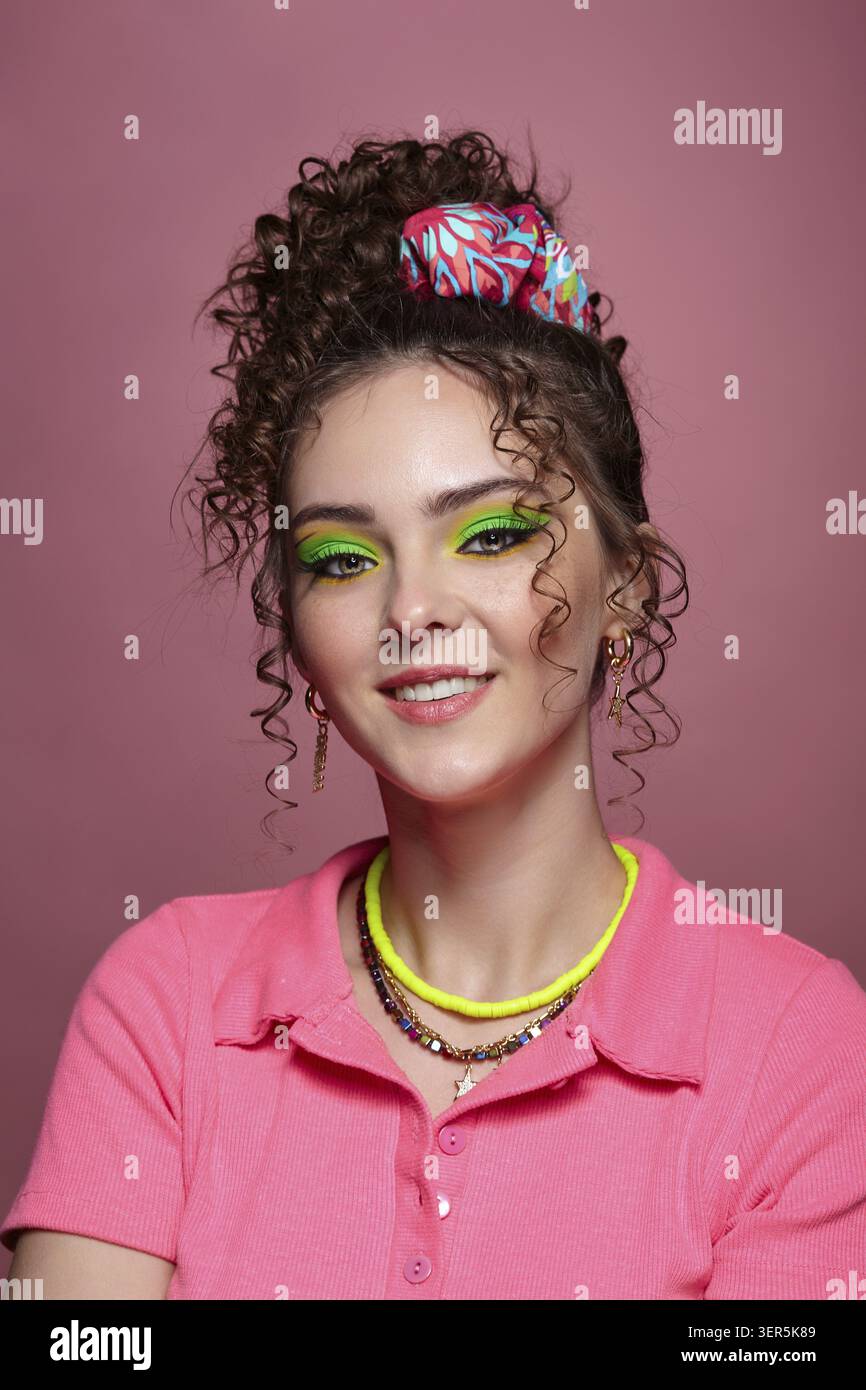 Ritratto di una giovane donna sorridente vestita con un abito rosa su sfondo rosa. Donna con insoliti occhi verdi ombre trucco, capelli ricci e orecchini Foto Stock