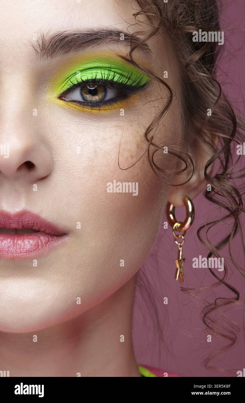Ritratto di una giovane donna su sfondo rosa. Donna con insoliti occhi verdi ombre trucco, capelli ricci e orecchini Foto Stock