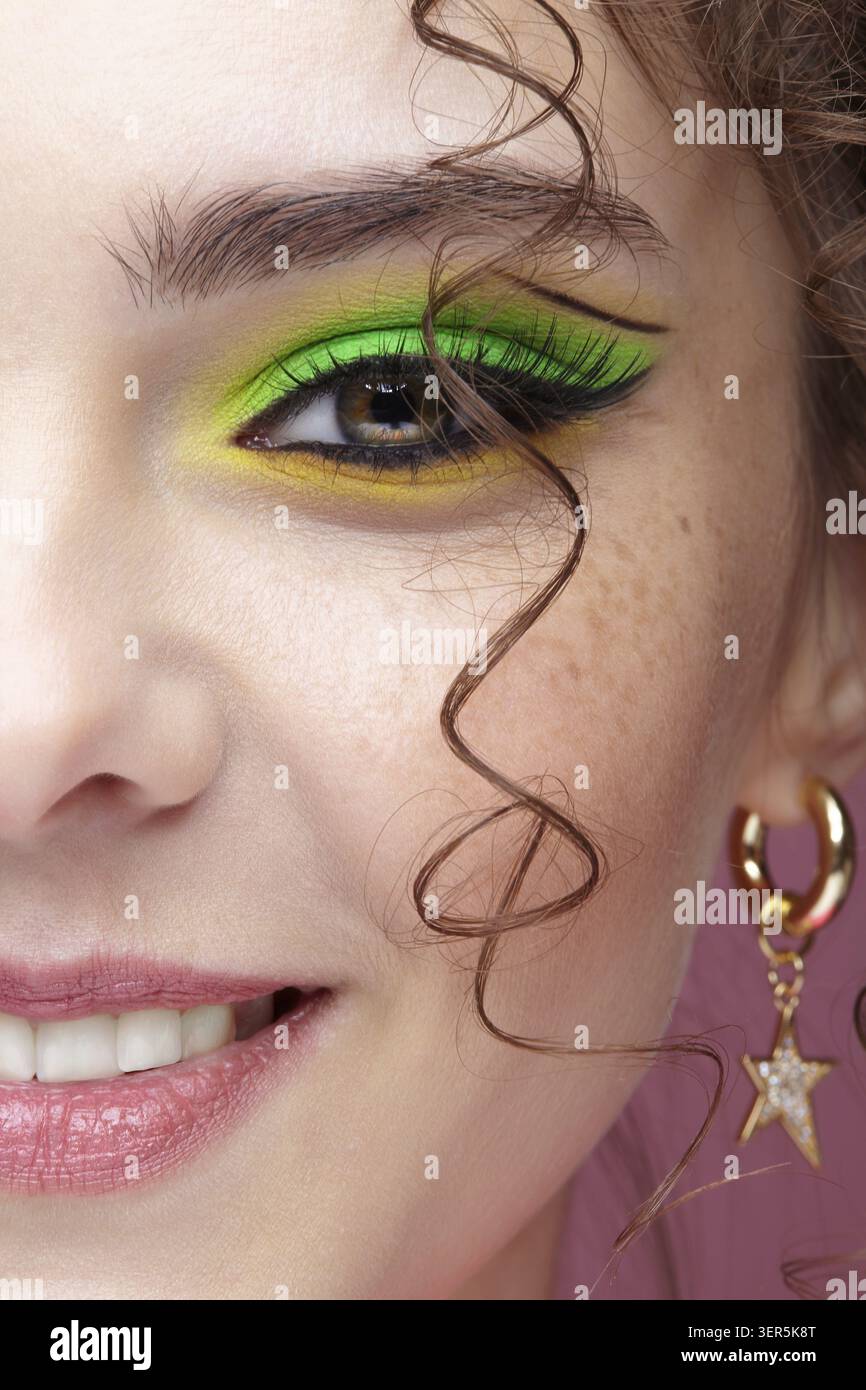 Ritratto di una giovane donna sorridente su sfondo rosa. Donna con insoliti occhi verdi ombre trucco, capelli ricci e orecchini Foto Stock