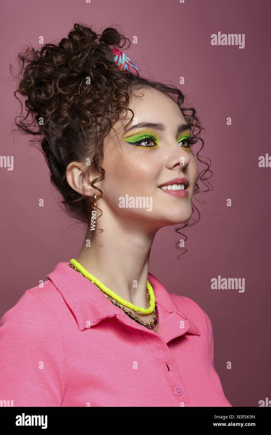 Ritratto di una giovane donna sorridente vestita con un abito rosa su sfondo rosa. Donna con insoliti occhi verdi ombre trucco, capelli ricci e orecchini Foto Stock