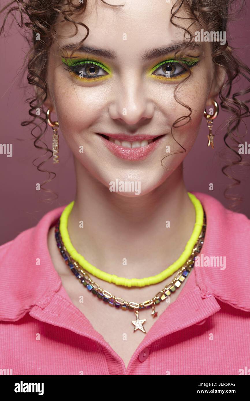 Ritratto di una giovane donna sorridente vestita con un abito rosa su sfondo rosa. Donna con insoliti occhi verdi ombre trucco, capelli ricci e orecchini Foto Stock
