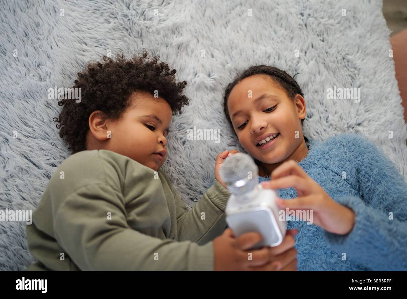 Due bambini sono felicemente impegnati in un'attività divertente su una morbida e soffice coperta. Il ragazzo guarda da vicino mentre la ragazza gli mostra come usare un piccolo gadget, Foto Stock