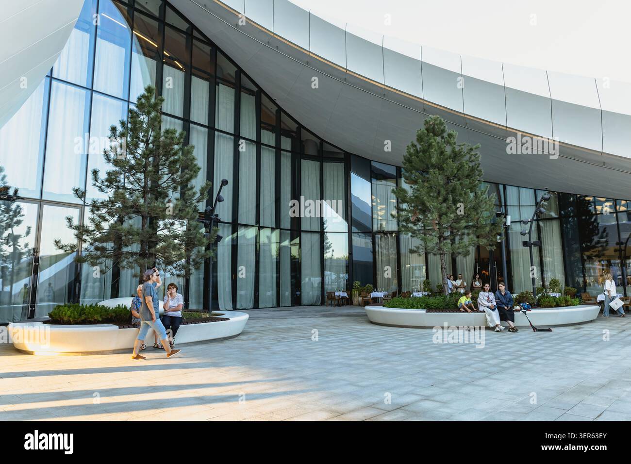Edificio contemporaneo con grandi finestre di vetro con tetto bianco curvo. Persone che camminano sedute su panchine bianche vicino ai pini. Gelendzhik, Russia - 1 giugno Foto Stock