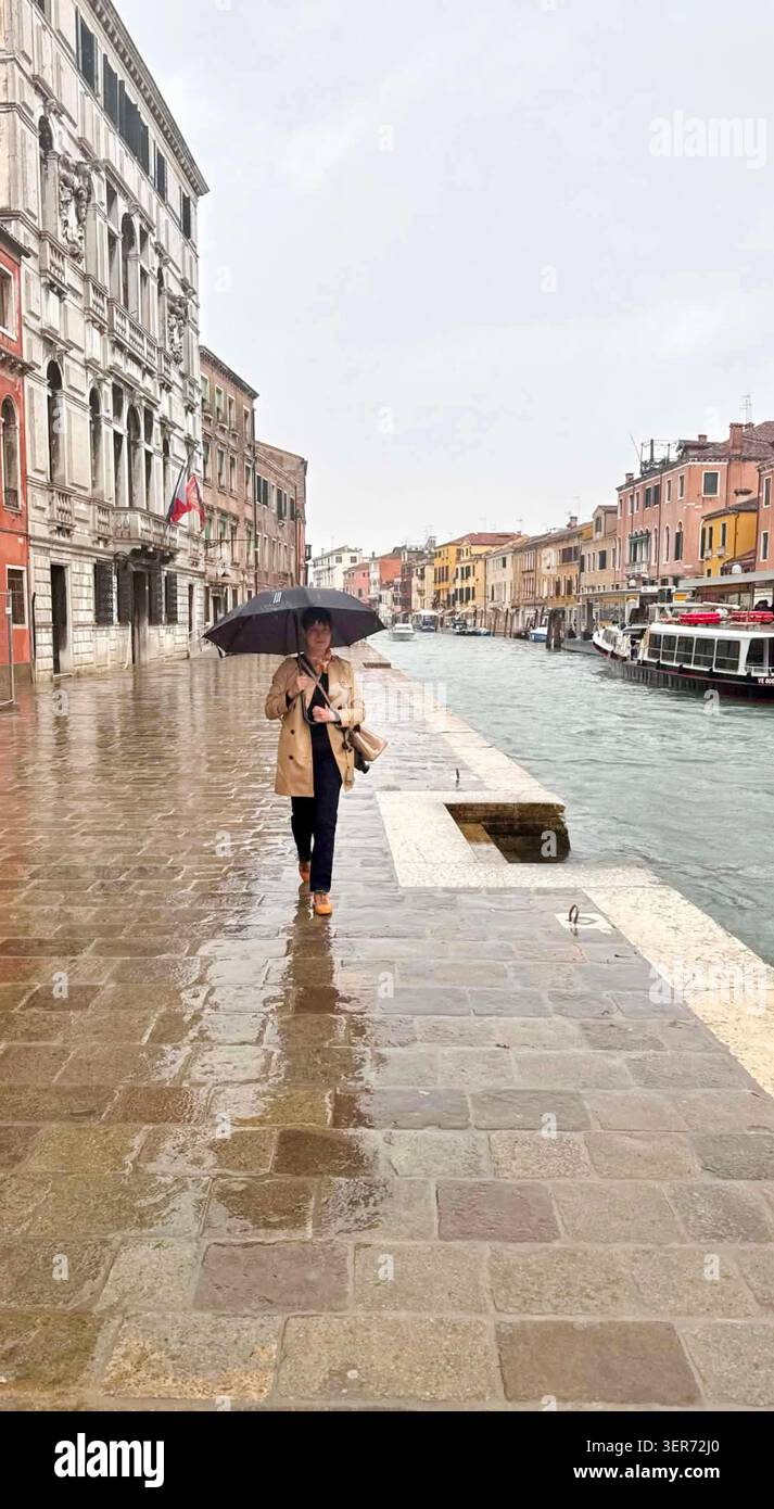 Donna che cammina con un ombrello accanto a un canale in una giornata di pioggia a Canareggio Venezia, Italia. Ha trent'anni e indossa un trench coat. Foto Stock