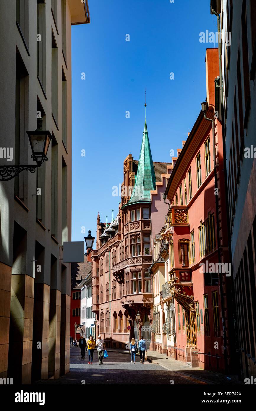 Vista della Haus zum Walfisch con la sua facciata in arenaria rossa ornata e una torretta dalle guglie verdi mentre la gente cammina lungo la strada di Friburgo in Breisgau, Foto Stock