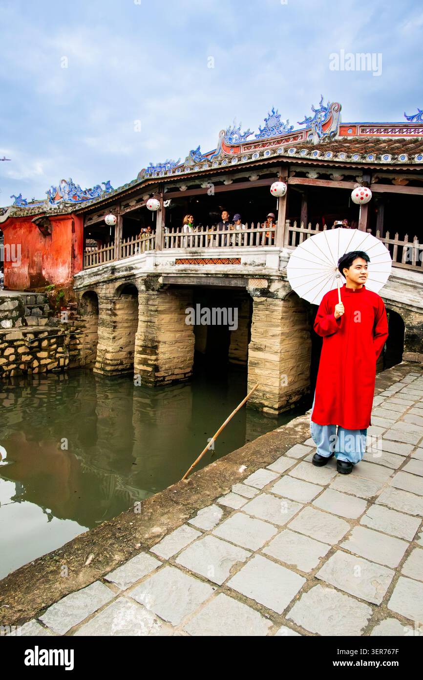Giovane uomo in abiti tradizionali davanti al ponte coperto giapponese storico a Hol An in Vietnam. Il ponte è stato costruito alla fine del XVI secolo Foto Stock