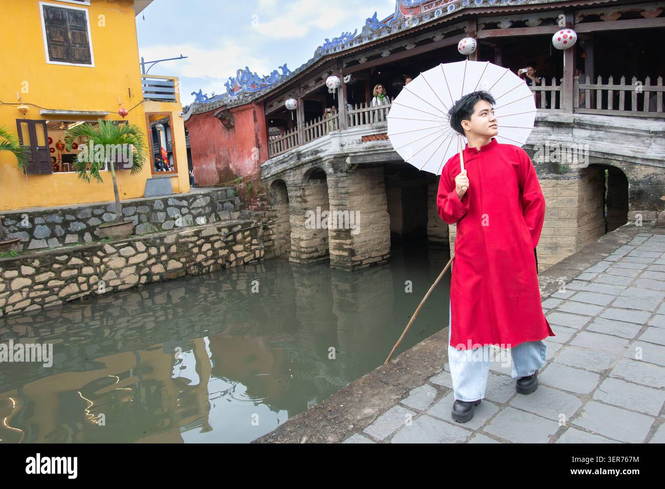 Un giovane con abiti tradizionali posa di fronte allo storico ponte coperto giapponese a HolAn Vietnam. Il ponte fu costruito alla fine del XVI secolo Foto Stock