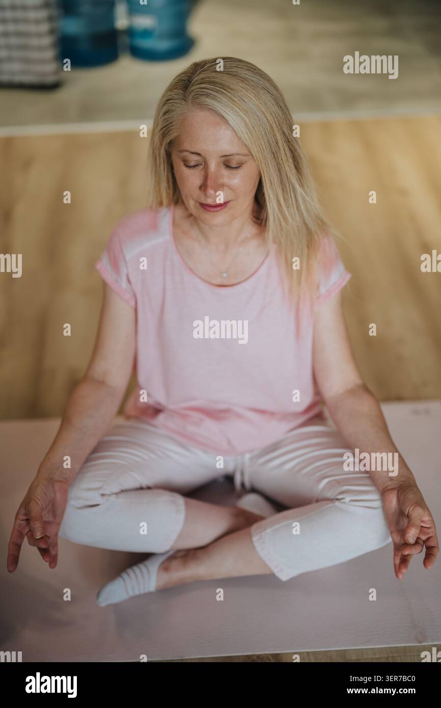 Donna matura che pratica meditazione e yoga seduta a gambe incrociate su un tappetino a casa Foto Stock