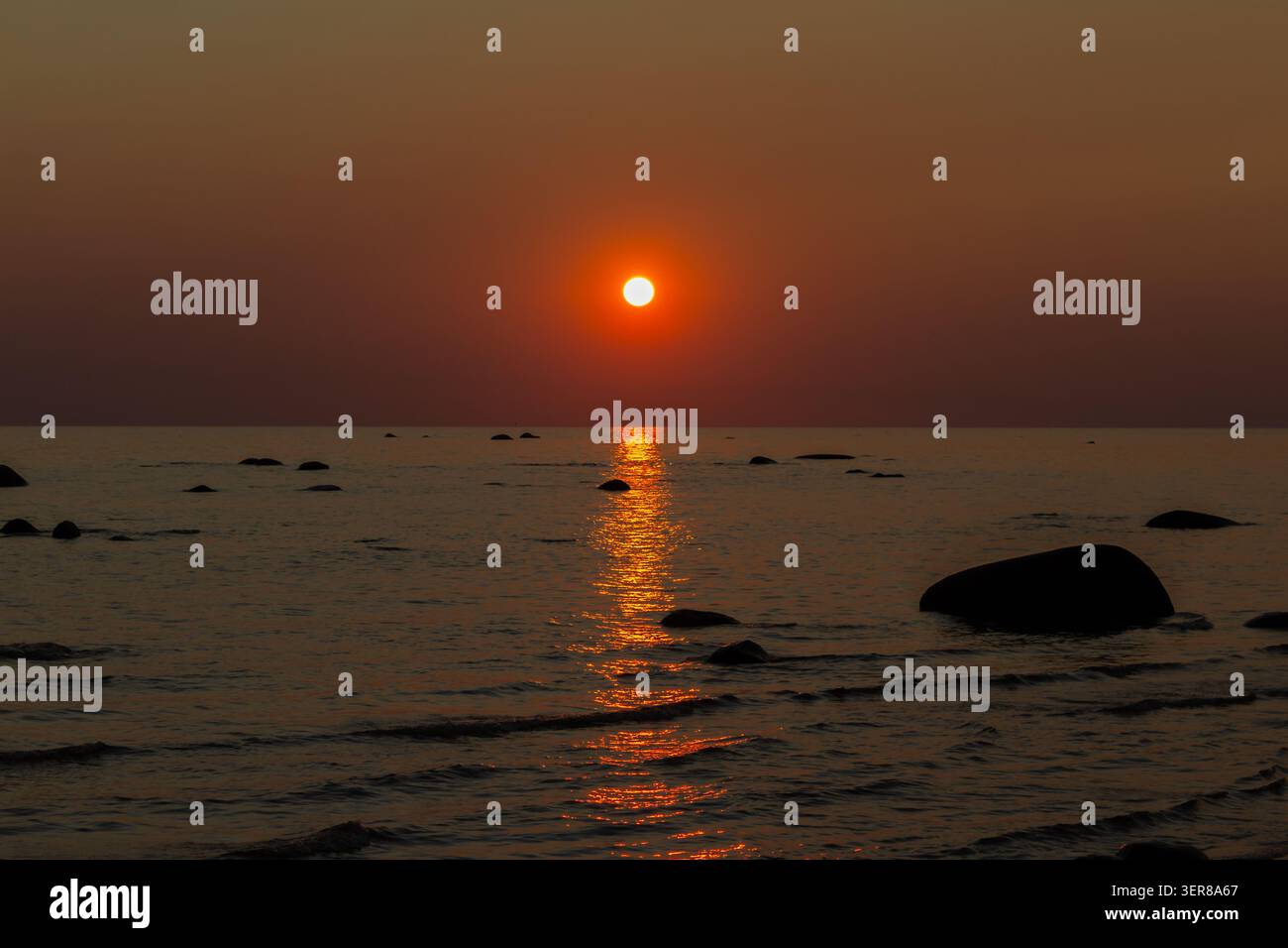 Tramonto rosso su un mare calmo con un riflesso solare brillante sull'acqua e scogliere costiere scure lungo la riva Foto Stock