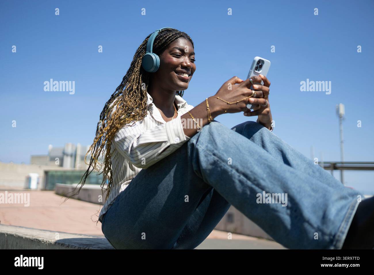 Una donna con i capelli intrecciati ama ascoltare musica con le cuffie wireless mentre è seduta all'aperto. Ha uno smartphone e sorride sotto una clea Foto Stock
