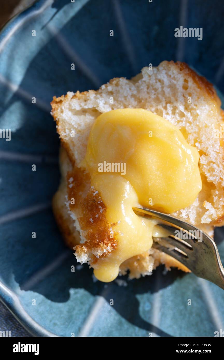 Foto ravvicinata della cagliata di limone dolce che gocciola su una torta di busto su un piatto blu, con una forchetta. Dessert agli agrumi, panini fatti in casa Foto Stock