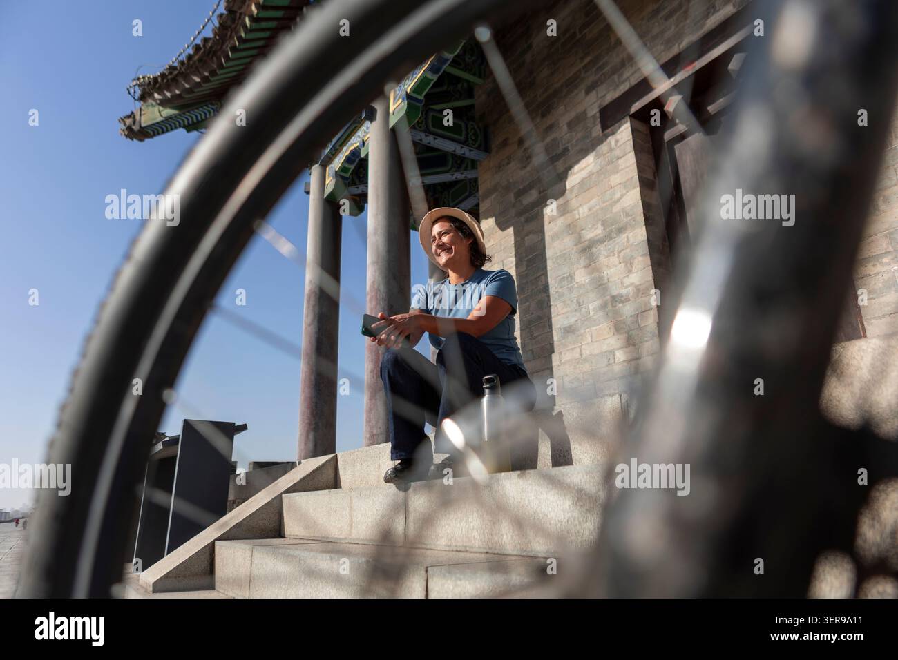 Una donna riposa in bicicletta accanto alle antiche mura della città di Xi'an, in Cina, incorniciata da raggi della bicicletta in una giornata di sole, mescolando viaggio, patrimonio culturale e u Foto Stock