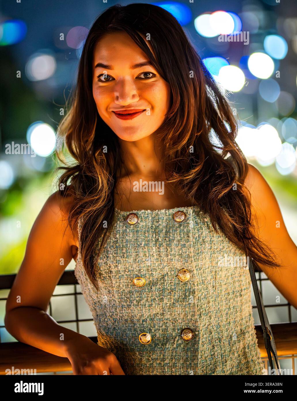 Giovane donna sorridente che posa all'aperto di notte con luci soffuse sullo sfondo. Illuminazione calda, abbigliamento informale e un'atmosfera rilassata e sicura creano la giusta atmosfera Foto Stock