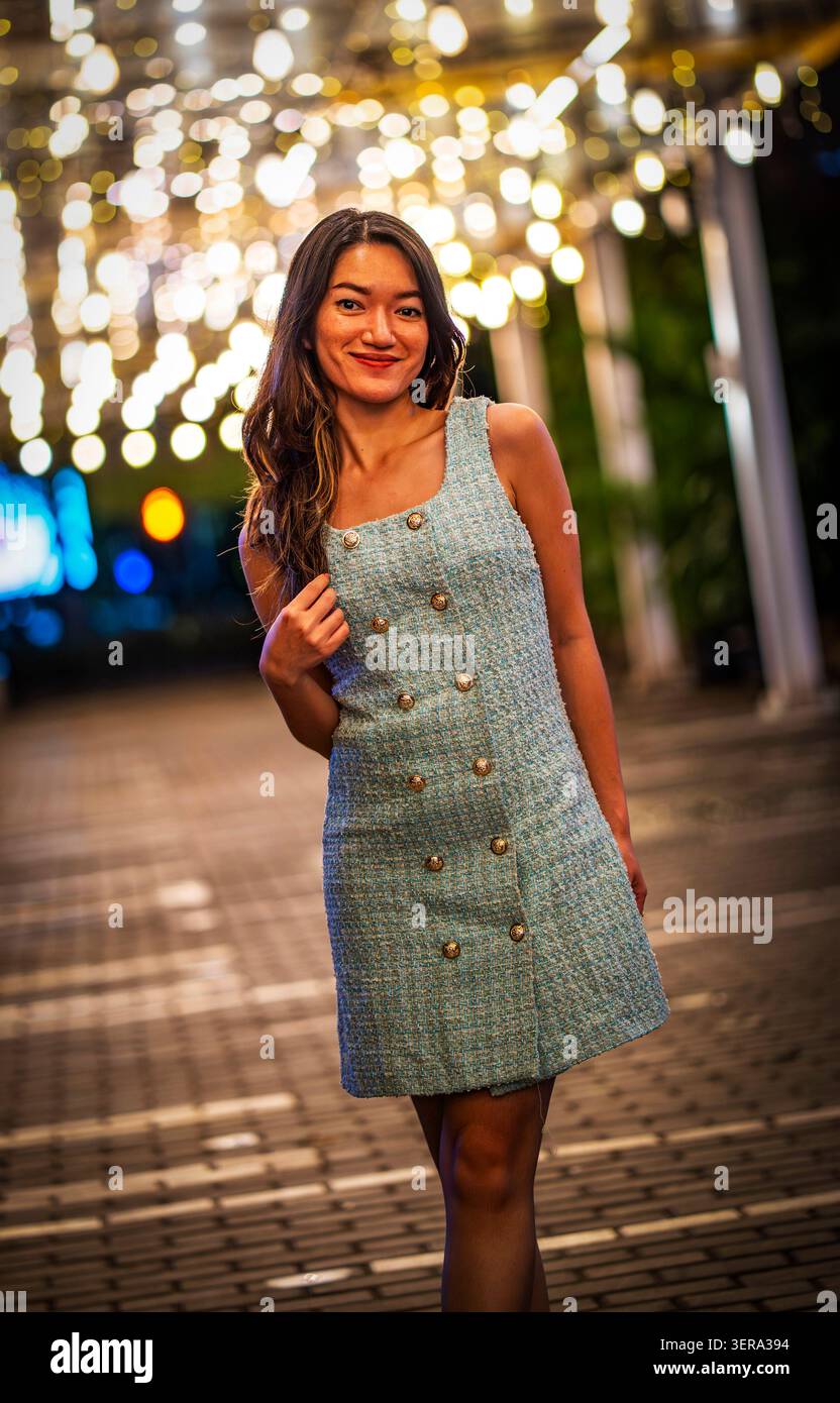 Giovane donna sorridente in un abito senza maniche che posa su un passaggio pedonale della città di notte, con calde luci a corda e morbido bokeh che creano una città elegante e allegra Foto Stock