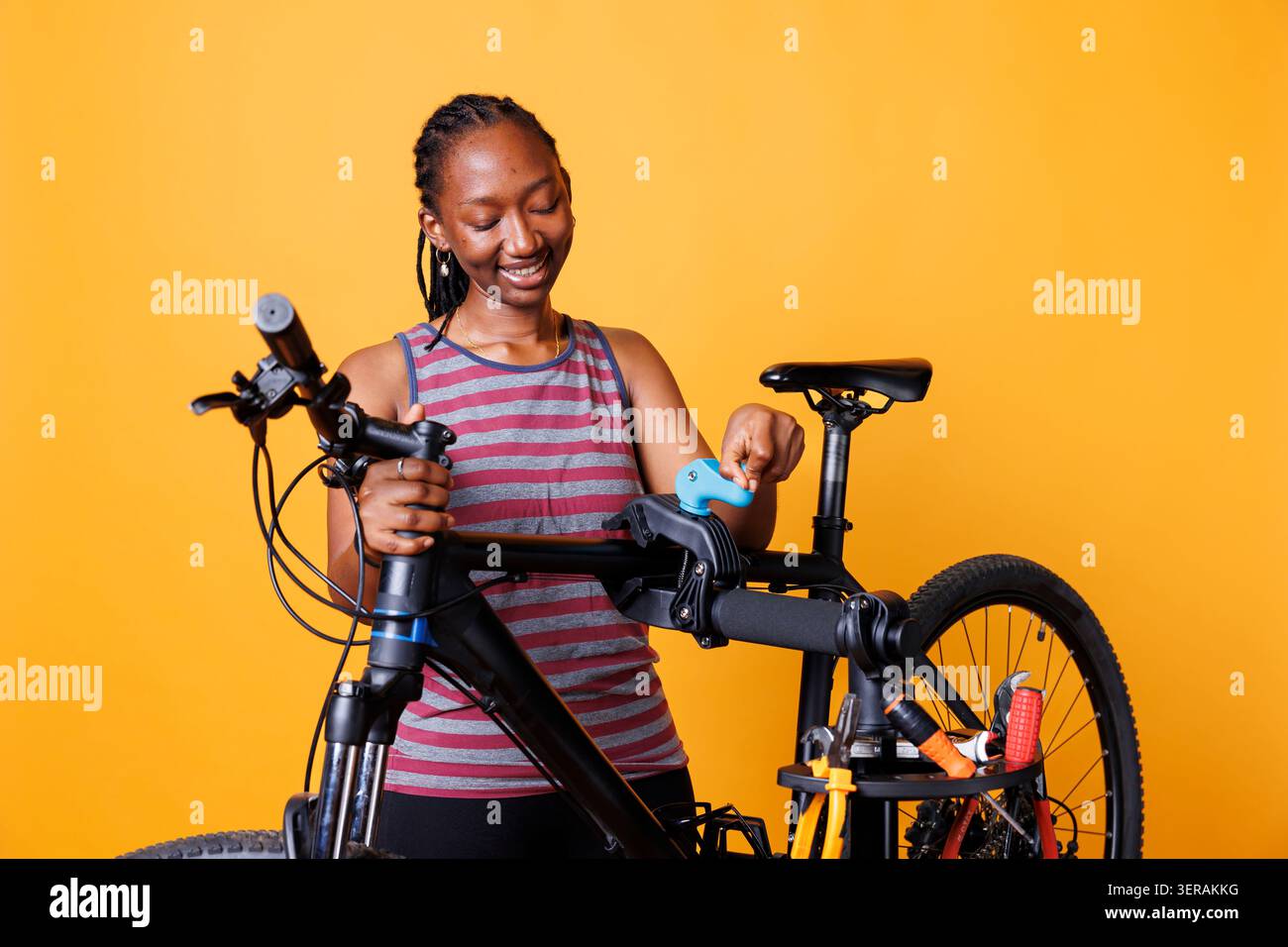 Donna energica che effettua regolazioni e ripara la moto con attrezzi speciali, con una donna dedicata che afferra ed esamina il telaio della bicicletta su un cavalletto di riparazione davanti a uno sfondo giallo. Foto Stock