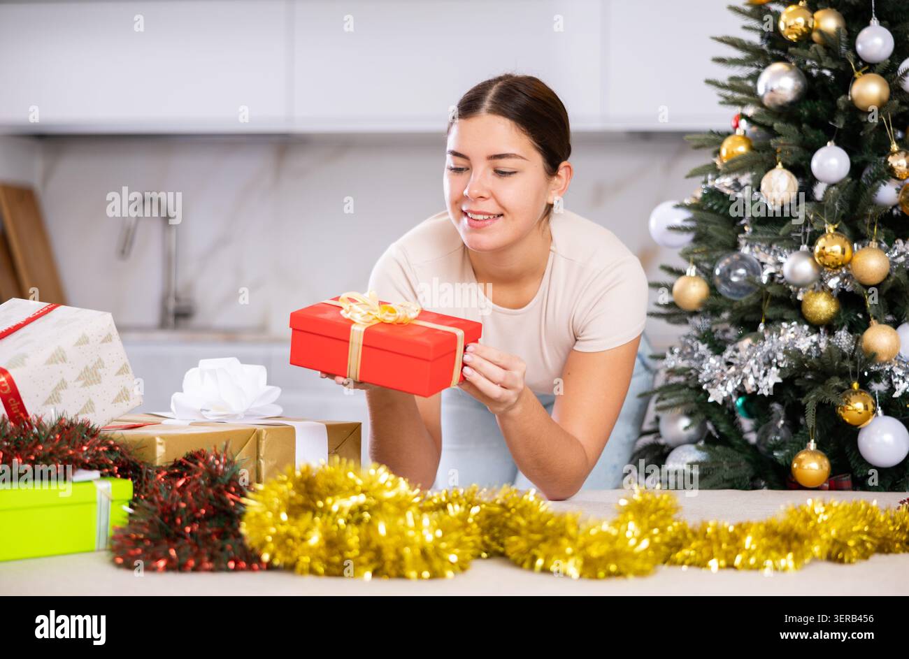 Giovane donna gioiosa in abiti casual che regge un regalo avvolto accanto all'albero di Natale in salotto a casa Foto Stock