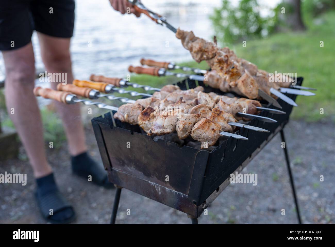 Una persona in piedi accanto a una griglia di metallo, tenendo uno spiedino di carne condita (shashlik) mentre cucina su carbone caldo in un parco panoramico sul lago. Questa immagine Foto Stock