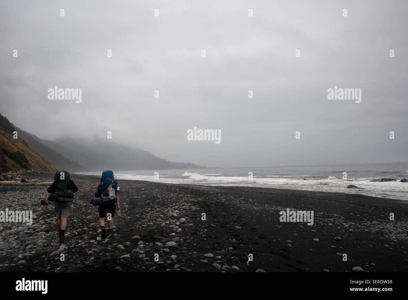 Vista di due escursionisti con grandi zaini che camminano lungo la spiaggia di sabbia nera con onde che si infrangono e montagne nebbiose sulla Lost Coast a Humboldt Count Foto Stock