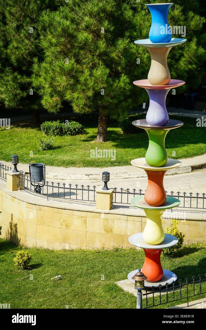 Una scultura all'aperto con sette bicchieri da tè e piattini Armudu stilizzati dai colori vivaci impilati verticalmente in un parco pubblico. Il backg Foto Stock