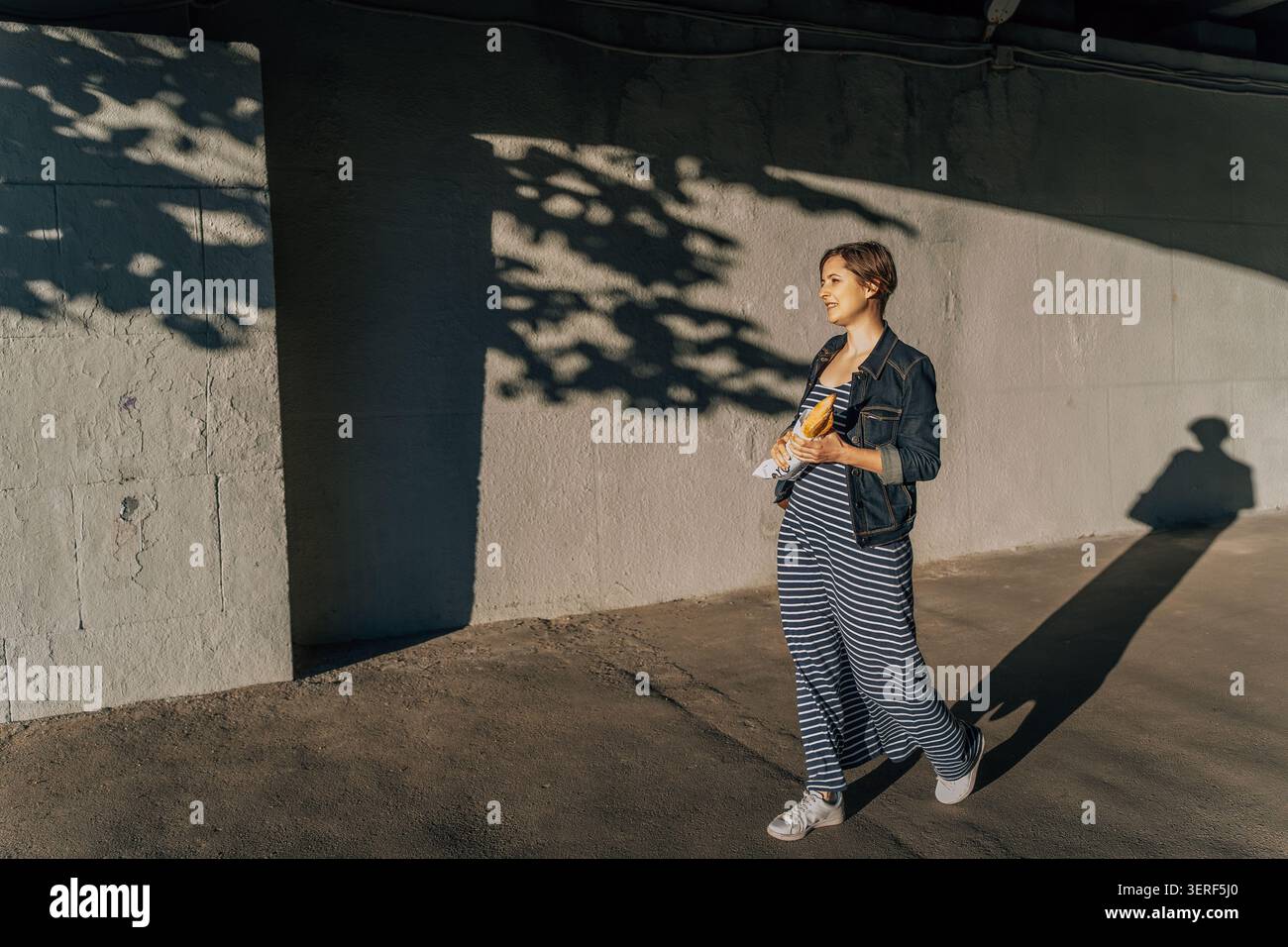 donna che cammina con la baguette alla luce del sole e getta lunghe ombre su una parete di cemento Foto Stock