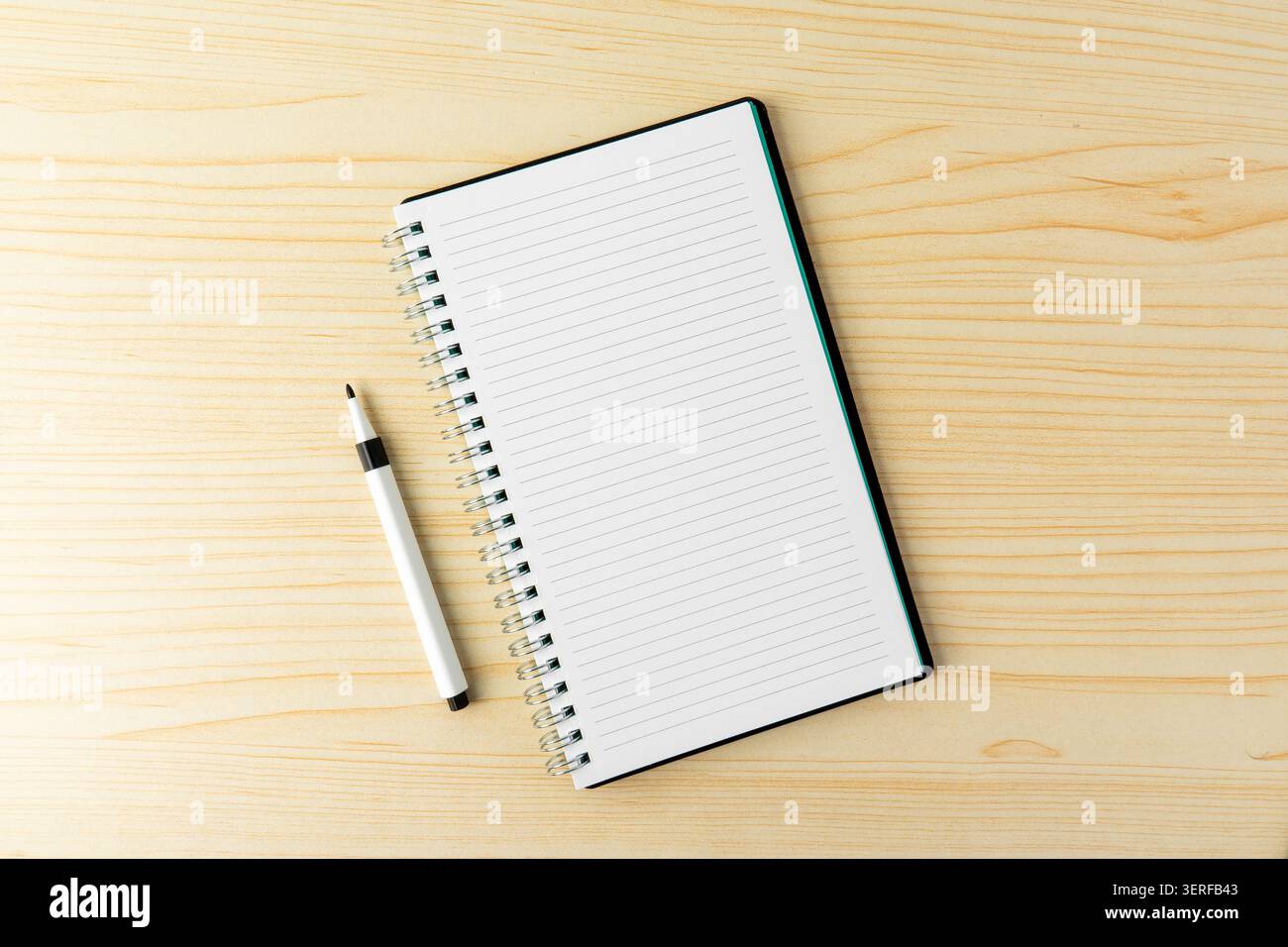 Un pennarello nero e un quaderno a spirale aperto rivestito in bianco sopra una scrivania di legno. Una composizione piatta minimalista che rappresenta la scrittura, l'educazione Foto Stock
