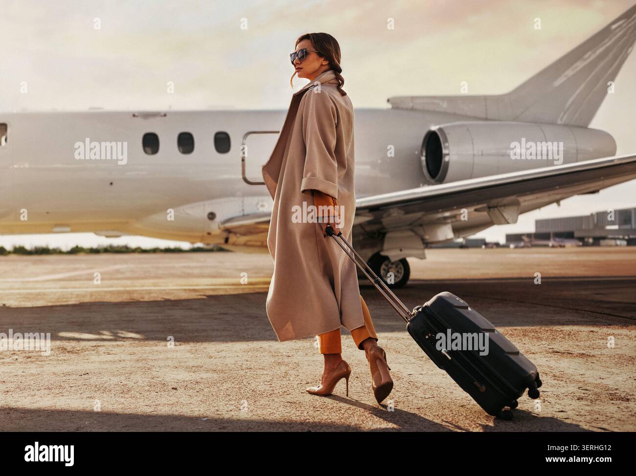 Elegante donna caucasica media adulta che cammina con valigia sulla pista dell'aeroporto con un jet privato Foto Stock