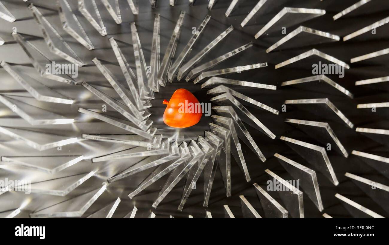 Vista dall'alto sull'installazione geometrica a spirale con elementi radiali metallici che circondano un oggetto rosso a forma di mela al centro. Forte simmetria, profondità Foto Stock