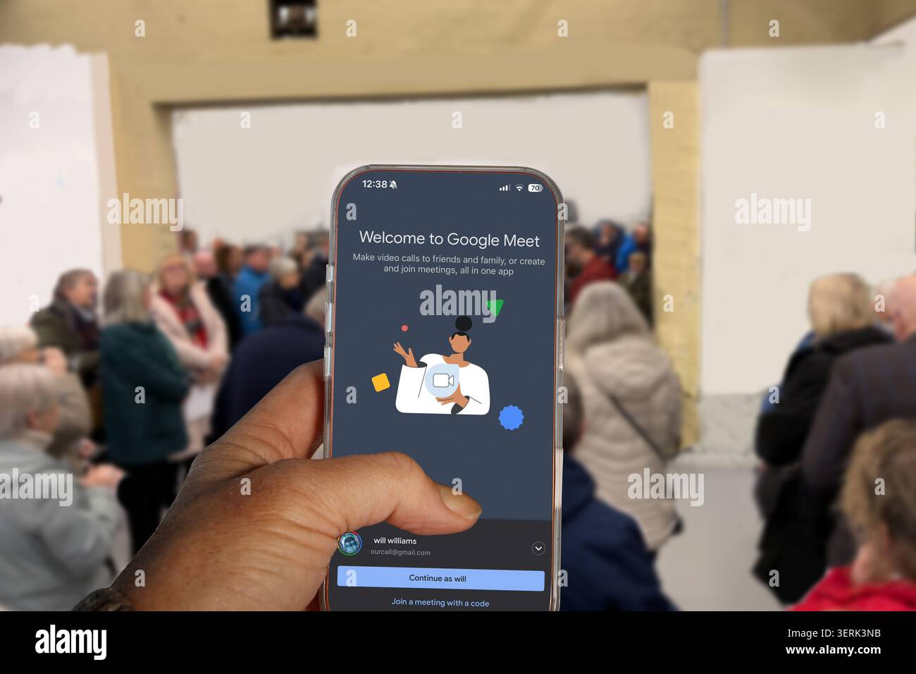 Mano che tiene in mano uno smartphone che visualizza l'app Google Meet con una folla sfocata di persone e spettatori sullo sfondo Foto Stock