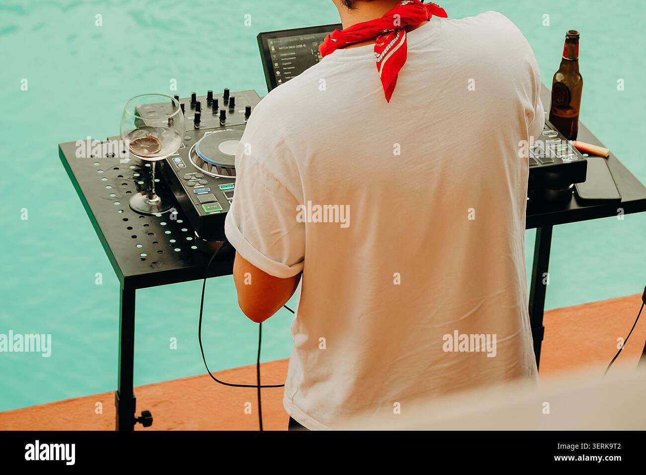 Il DJ mescola musica a bordo piscina durante un raduno estivo al sole del pomeriggio con un drink sul tavolo. Bicchieri e una bottiglia di birra sono nelle vicinanze come He pla Foto Stock