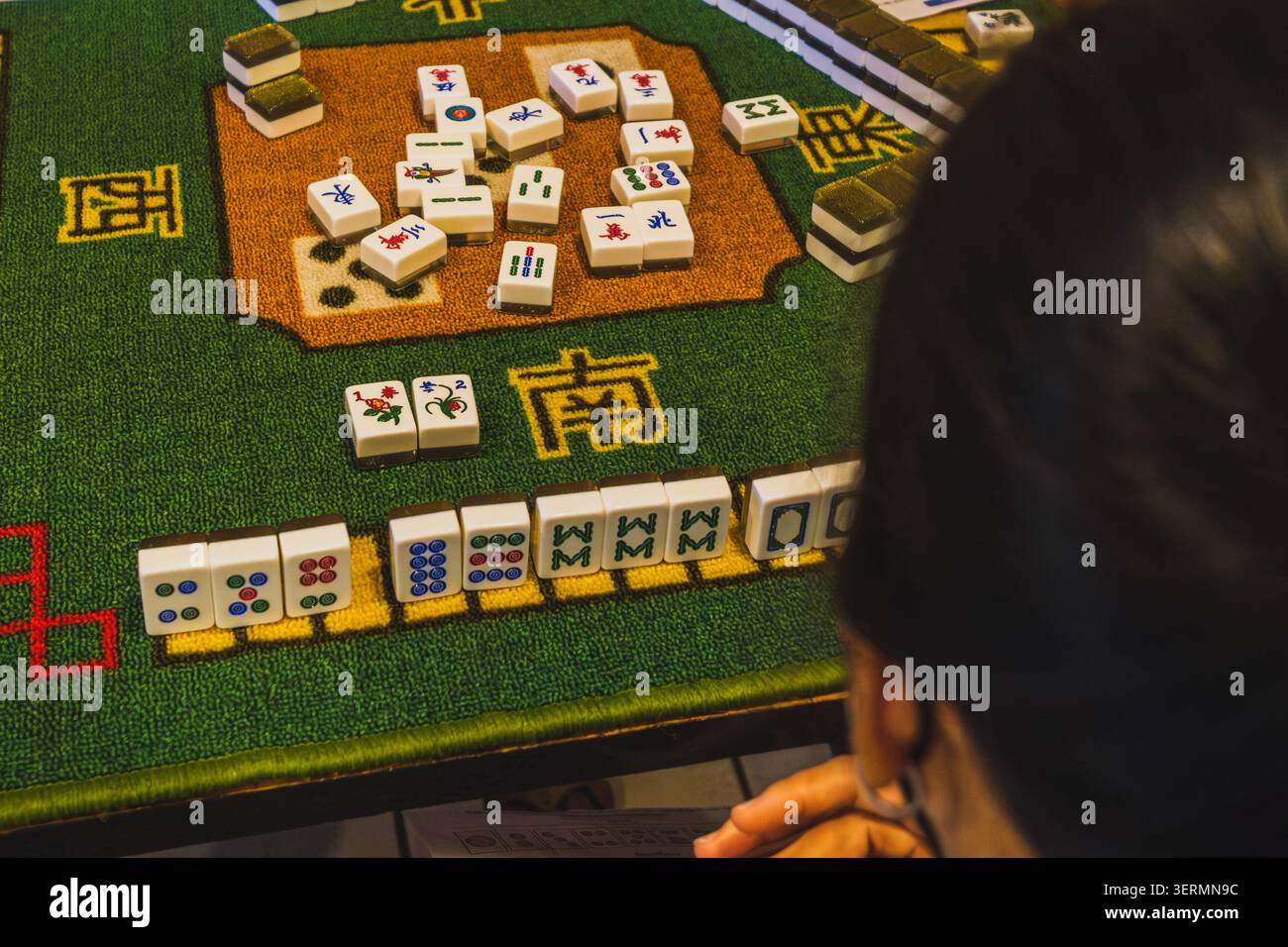 Una vista a spalla di una persona che gioca a Mahjong, mostrando una mano di tessere allineate su un tappetino verde, tessere scartate al centro e tessere floreali Foto Stock