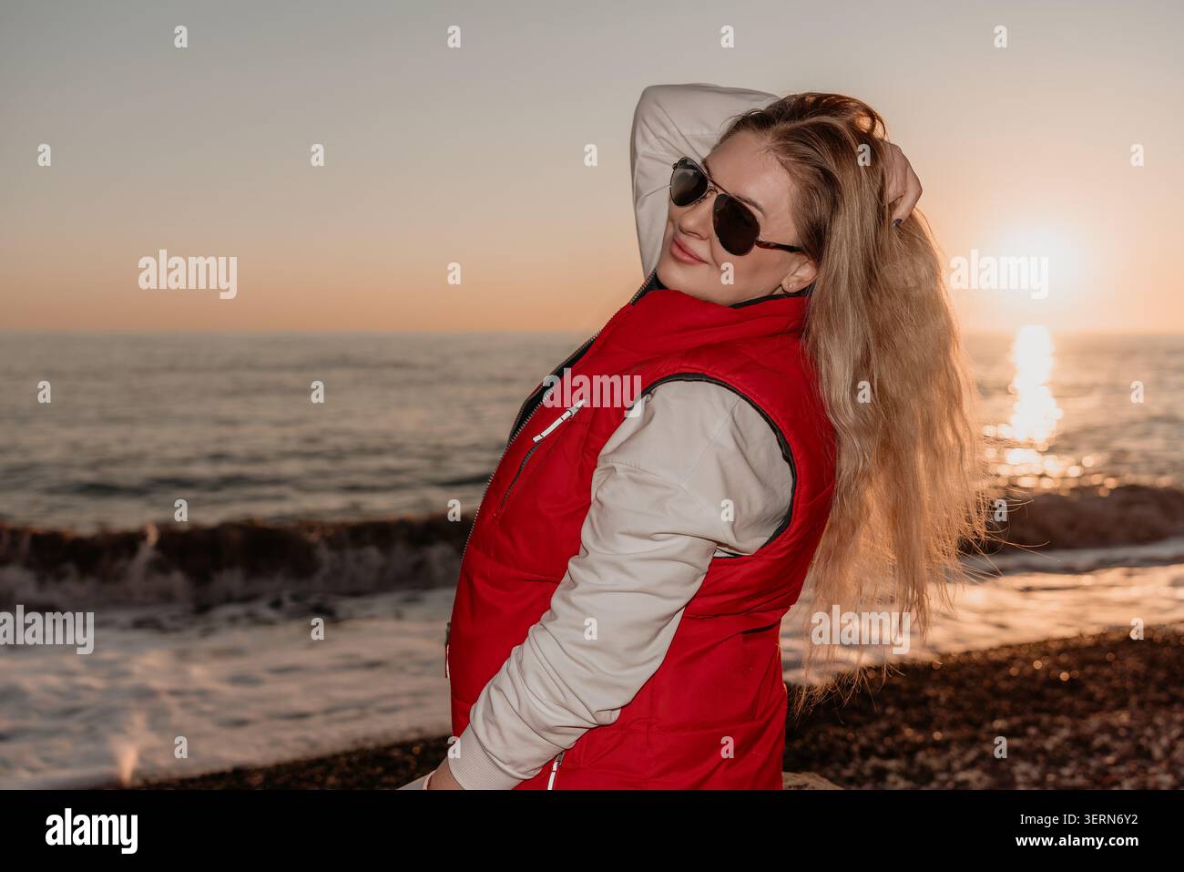 Donna al tramonto che posa in un gilet rosso e occhiali da sole durante l'ora d'oro su una riva costiera Foto Stock