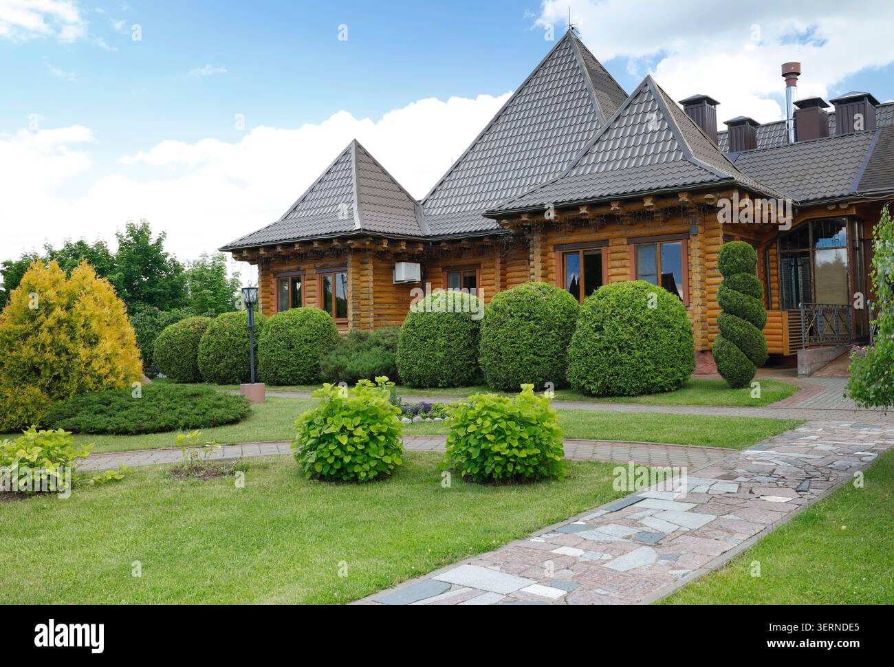 Bellissimi arbusti verdi curati e cespugli rotondi di fronte alla lussuosa villa in legno. Piante decorative sempreverdi con albero a spirale e sferico Foto Stock